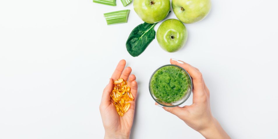 Top view female’s hands holding glass of green smoothie Top view female's hands holding glass of green smoothie and capsules of omega 3 nutritional supplements next to fresh apples, spinach and celery on white background, flat lay.