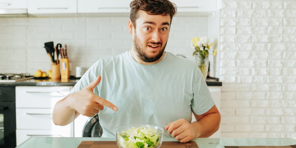 Young dieting man points finger bowl of salad in front of him asking Is this supposed to be eaten A young dieting man points his index finger at a bowl of salad in front of him, asking Is this supposed to be eaten Green salad healthy food concept. Sits in his kitchen. Looking at camera.